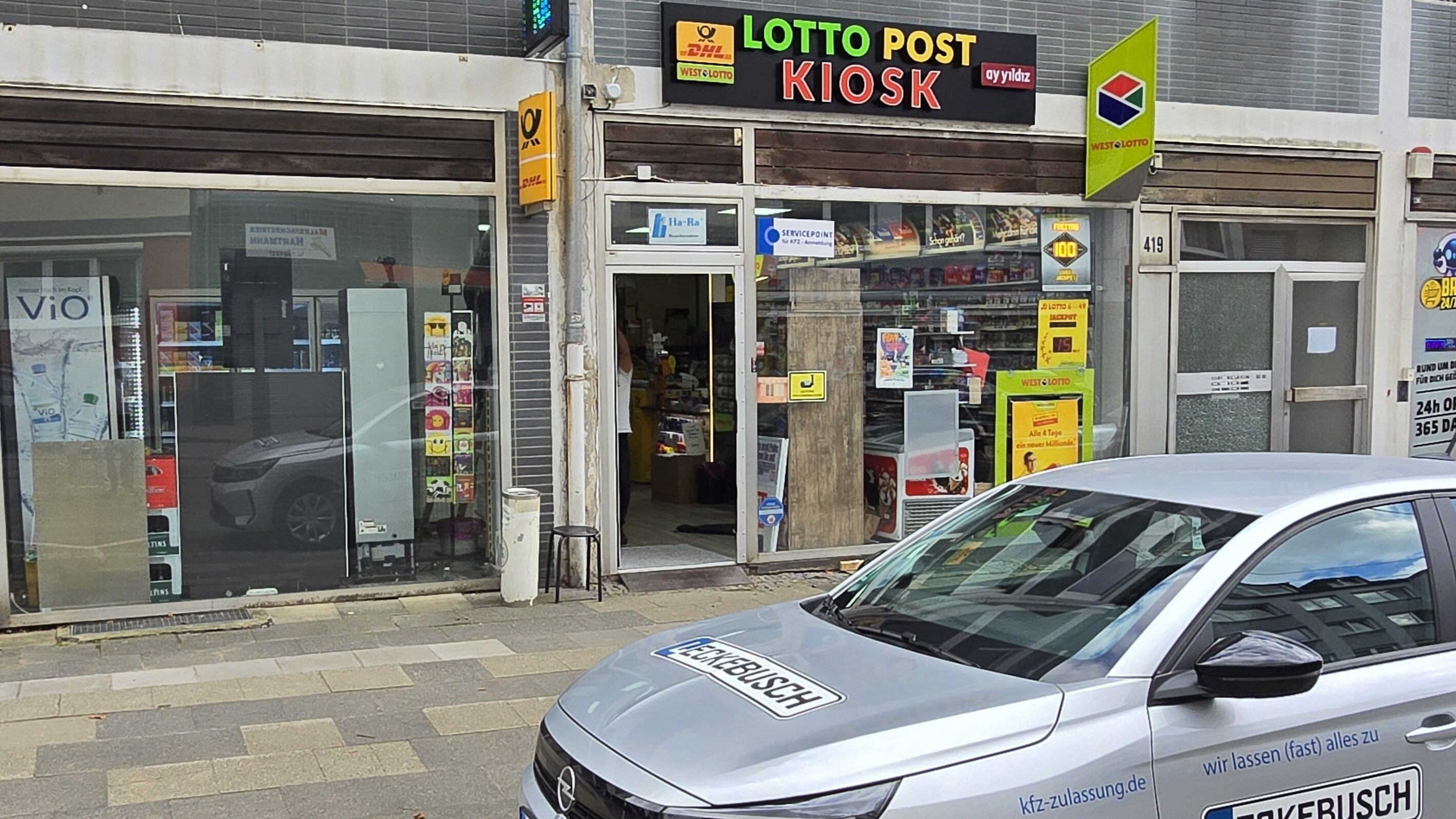 Ein graues Auto parkt vor einem Kiosk mit buntem Lotto-Schild.