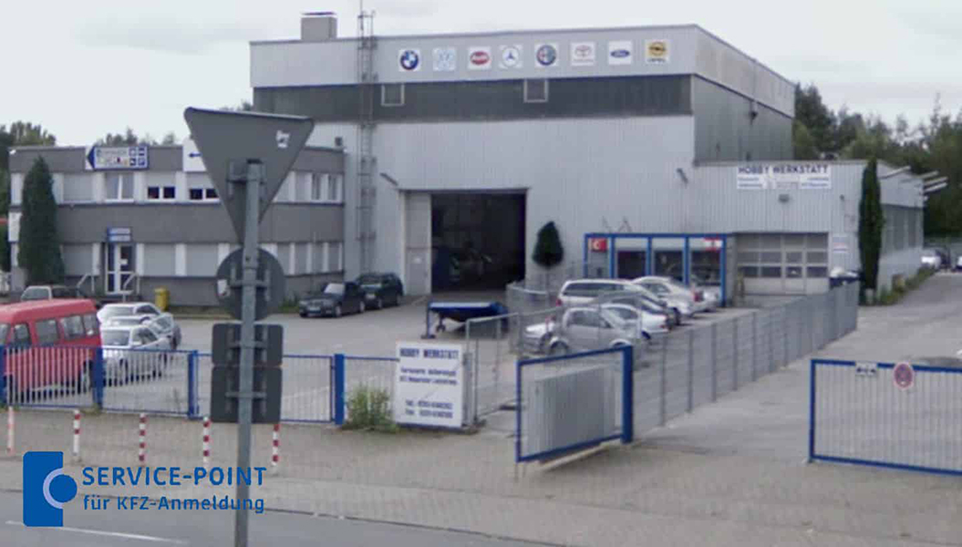 Großes silbernes Autohaus mit vielen Logos, mehreren geparkten Autos und blauem Zaun