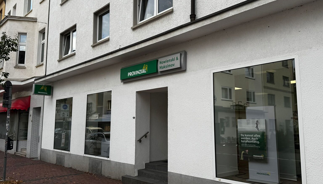 Das Bild zeigt eine weiße Fassade einer Versicherungsagentur mit großen Fenstern und grünen Schildern