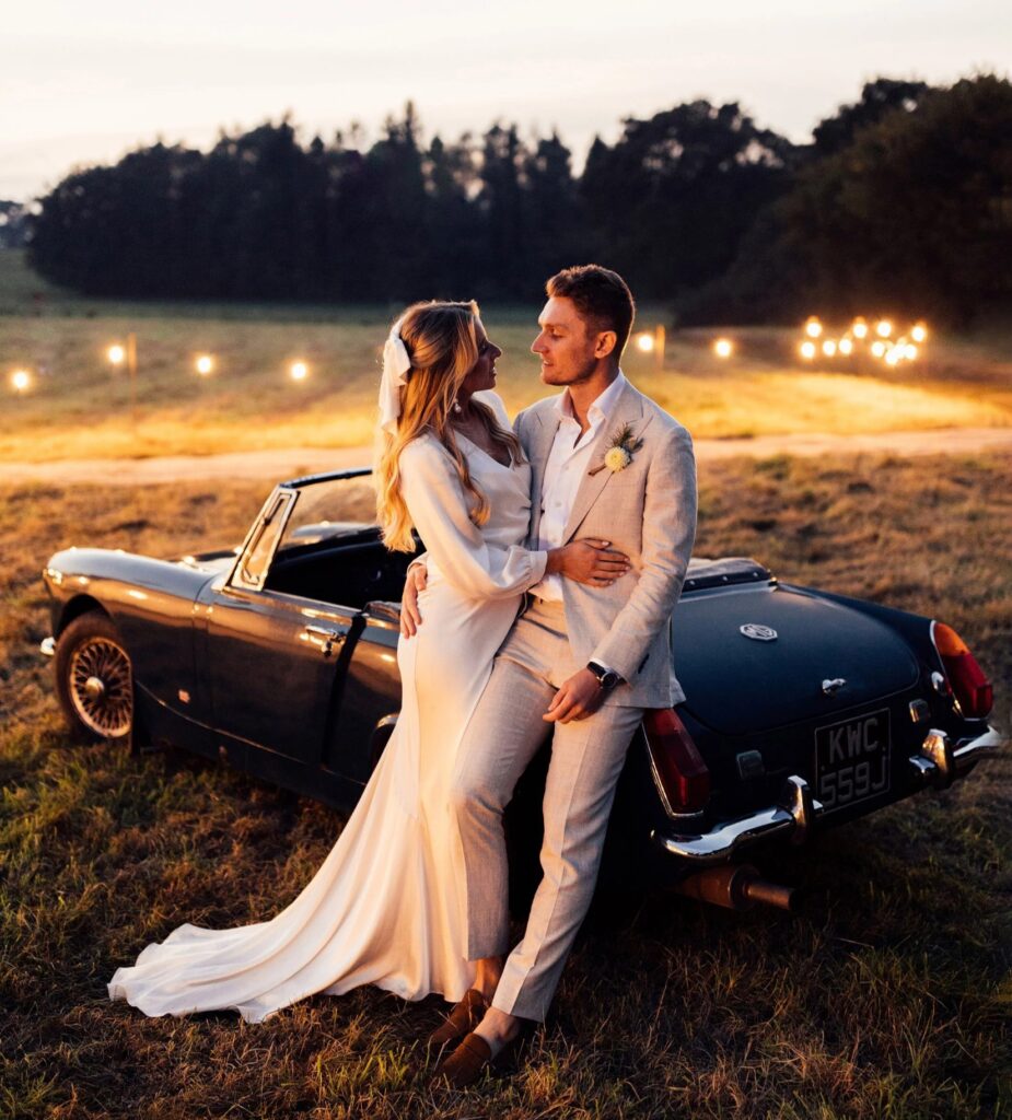 Ein Paar in festlicher Kleidung steht lächelnd vor einem dunklen Oldtimer im Abendlicht