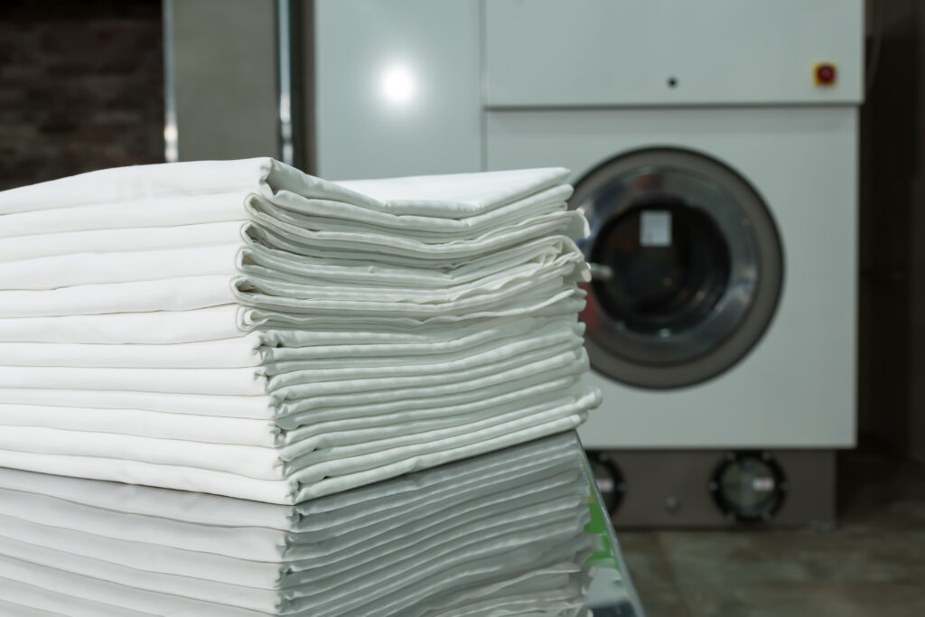 Ein Stapel frisch gefalteter weisser Bettwaesche liegt vor einer Industriewaschmaschine