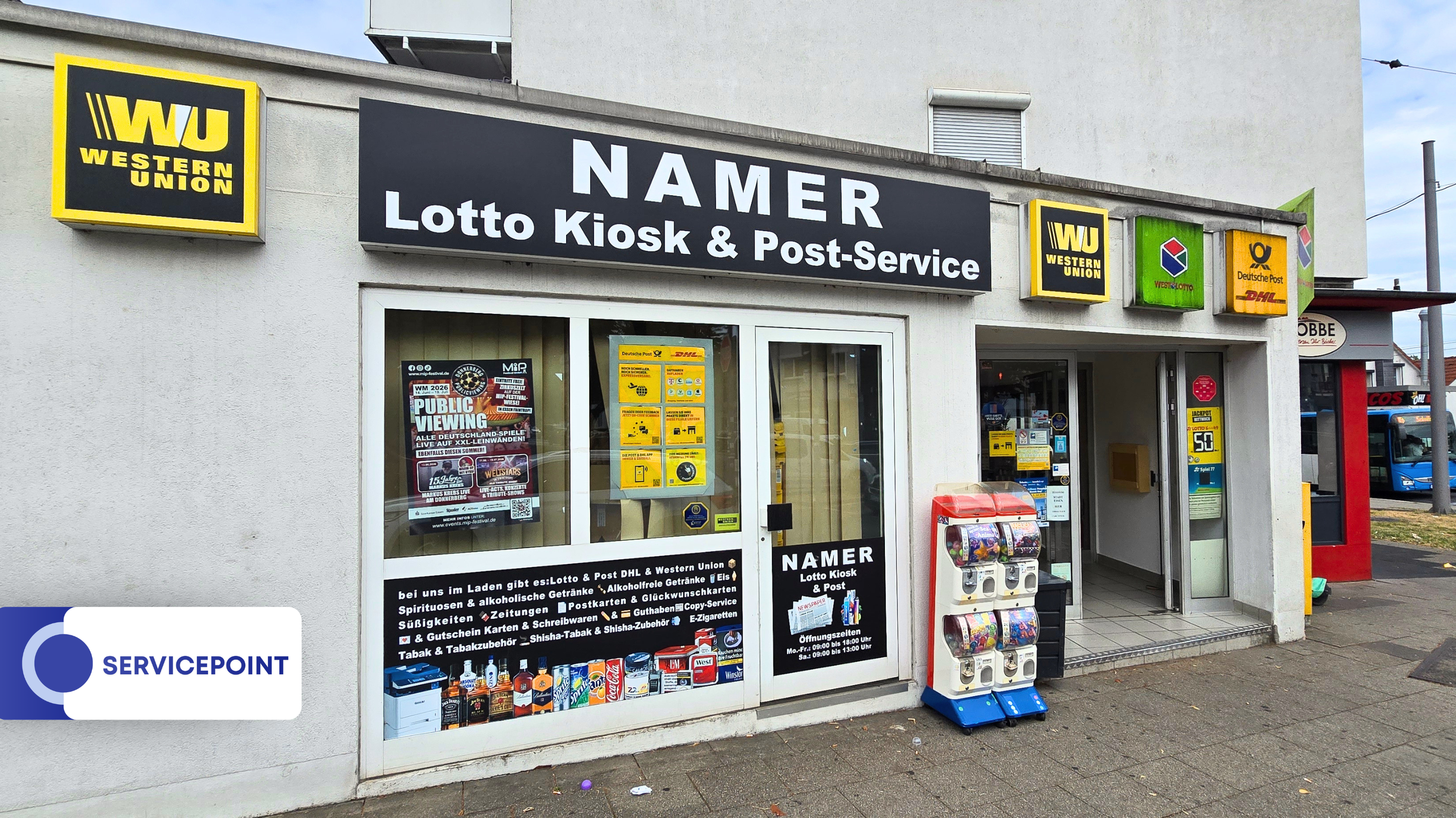 Lottokiosk Namer mit Schaufenster, gelben Schildern und Zeitungsständer vorne draußen